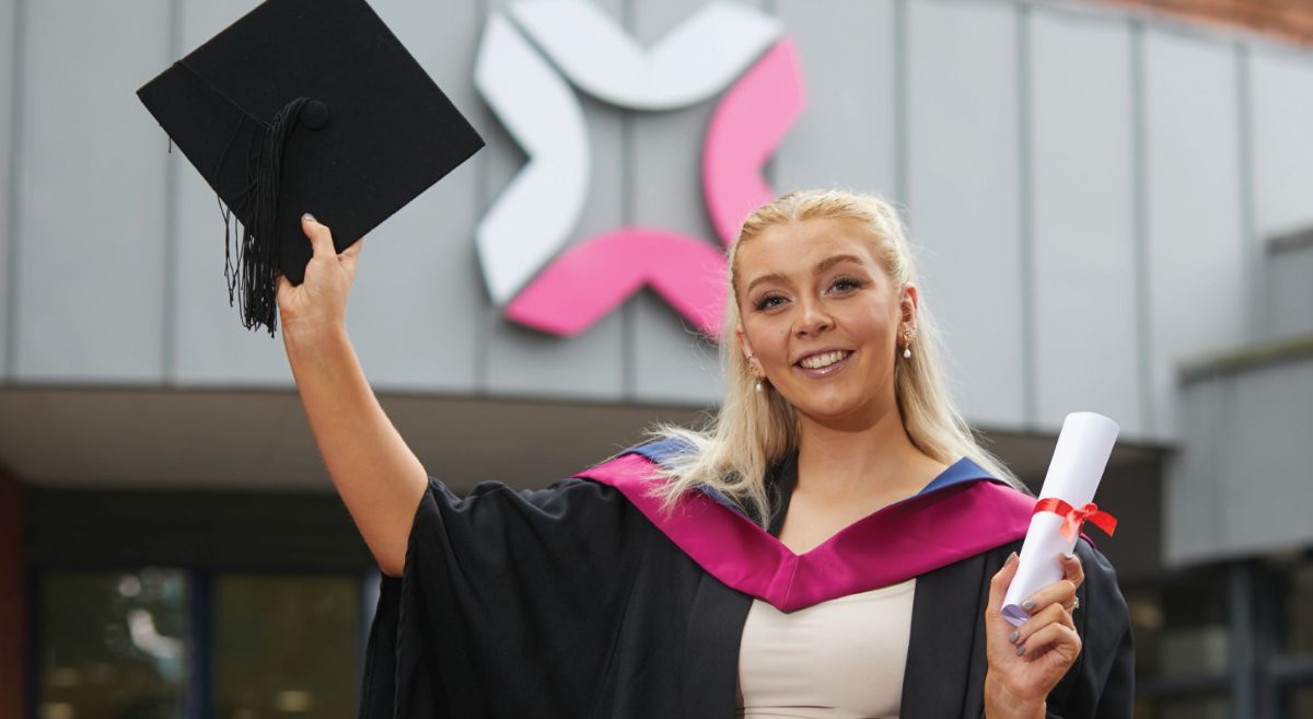 Image showing student Leah Hamilton standing outside Bangor Campus holding scroll and motorboard